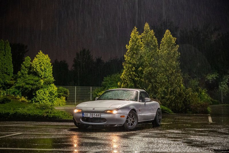 A Mazda Miata parked under the rain at night, with greenery in a Polish parking lot.