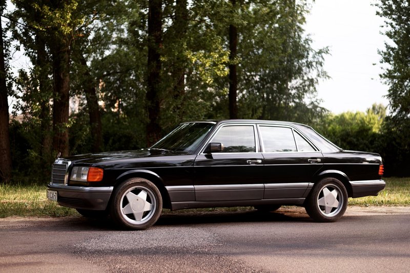 Vintage Mercedes W126 sedan parked by a tree-lined road on a sunny day.