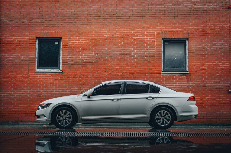 Side view of a silver sedan parked in front of a red brick wall with reflections on wet pavement.