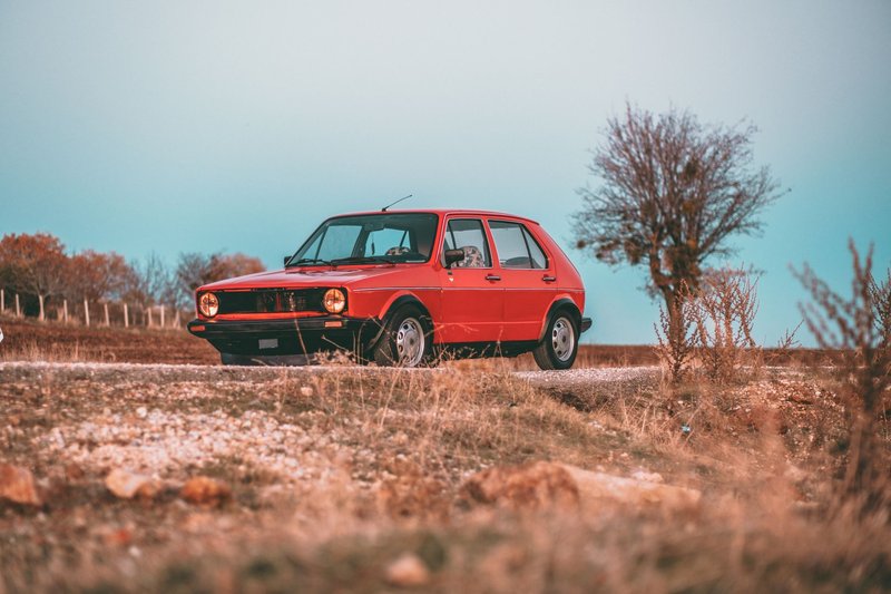 A classic red hatchback car parked on a rural dirt road against a natural backdrop.