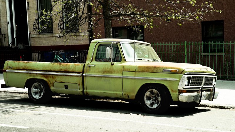 Rusty green vintage pickup truck parked on a city street with urban backdrop.
