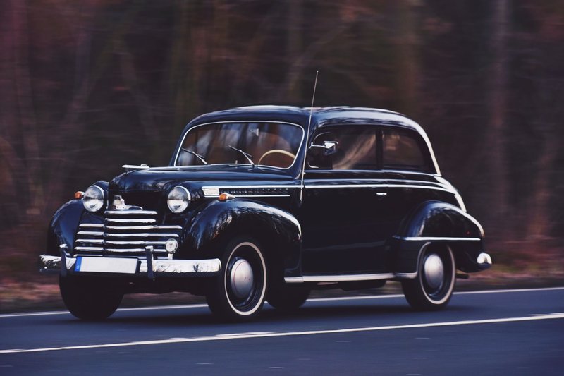 A sleek vintage car driving on a road with blurred wooded background, showcasing timeless elegance.