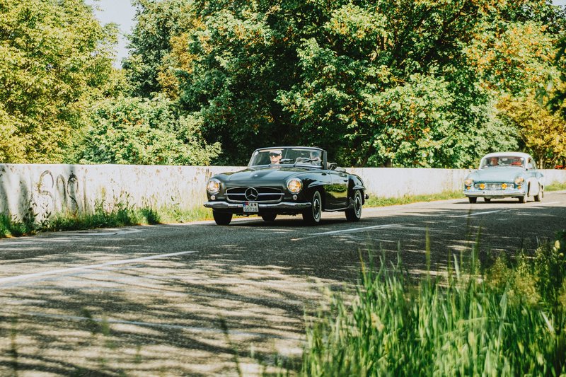 Two classic cars on a scenic country road surrounded by lush greenery in daylight.