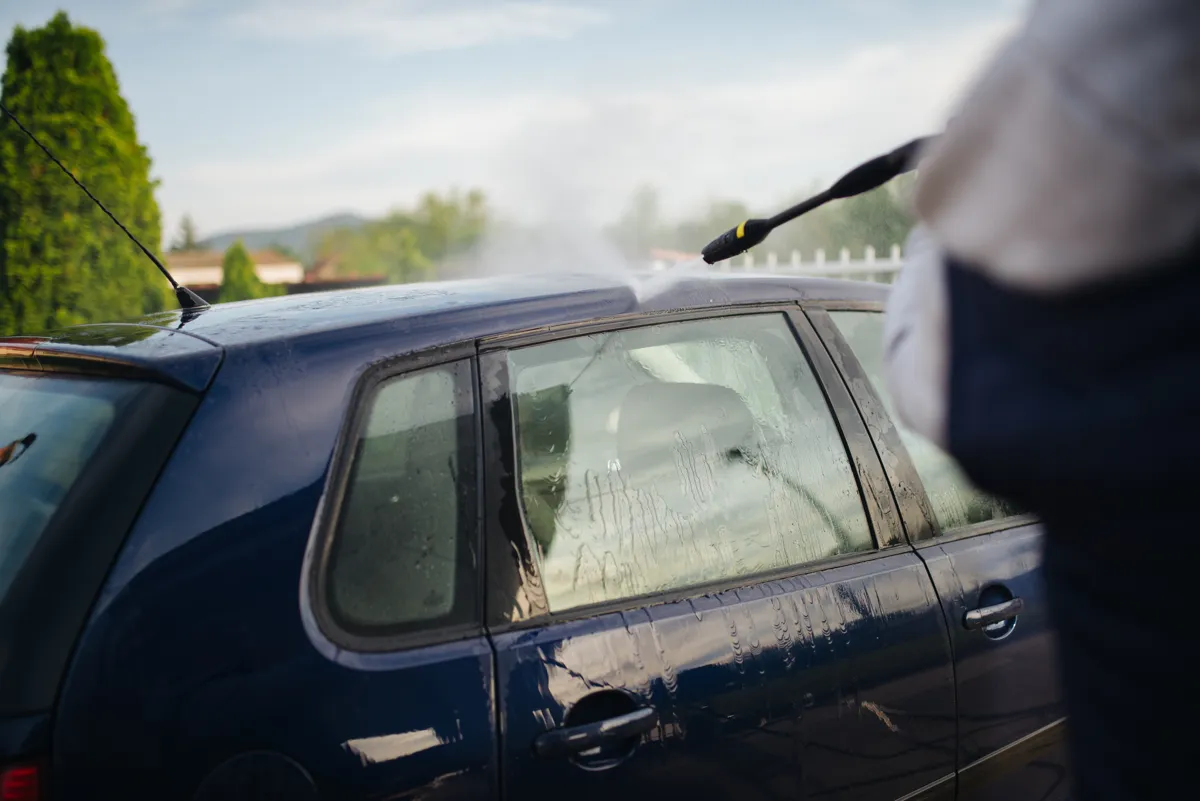 Person rinsing a dark blue hatchback with a pressure washer in a driveway